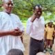 Two officials speak with pupils under a tree at a school compound