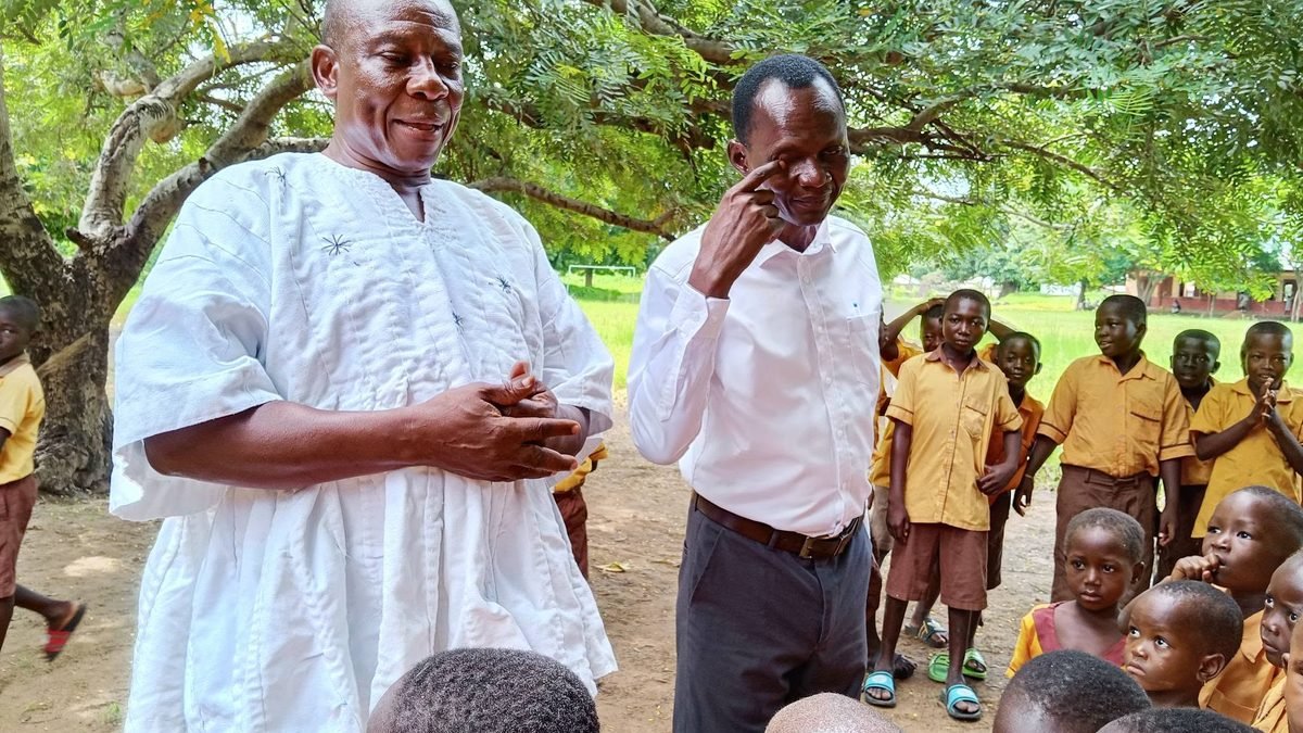 Two officials speak with pupils under a tree at a school compound